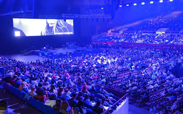 Large blue-lit arena packed with seated crowd facing a giant stage screen