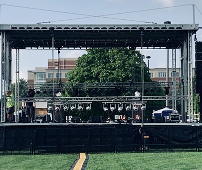 Outdoor stage with black backdrop, lighting rig, and people beneath a large canopy in a grassy field