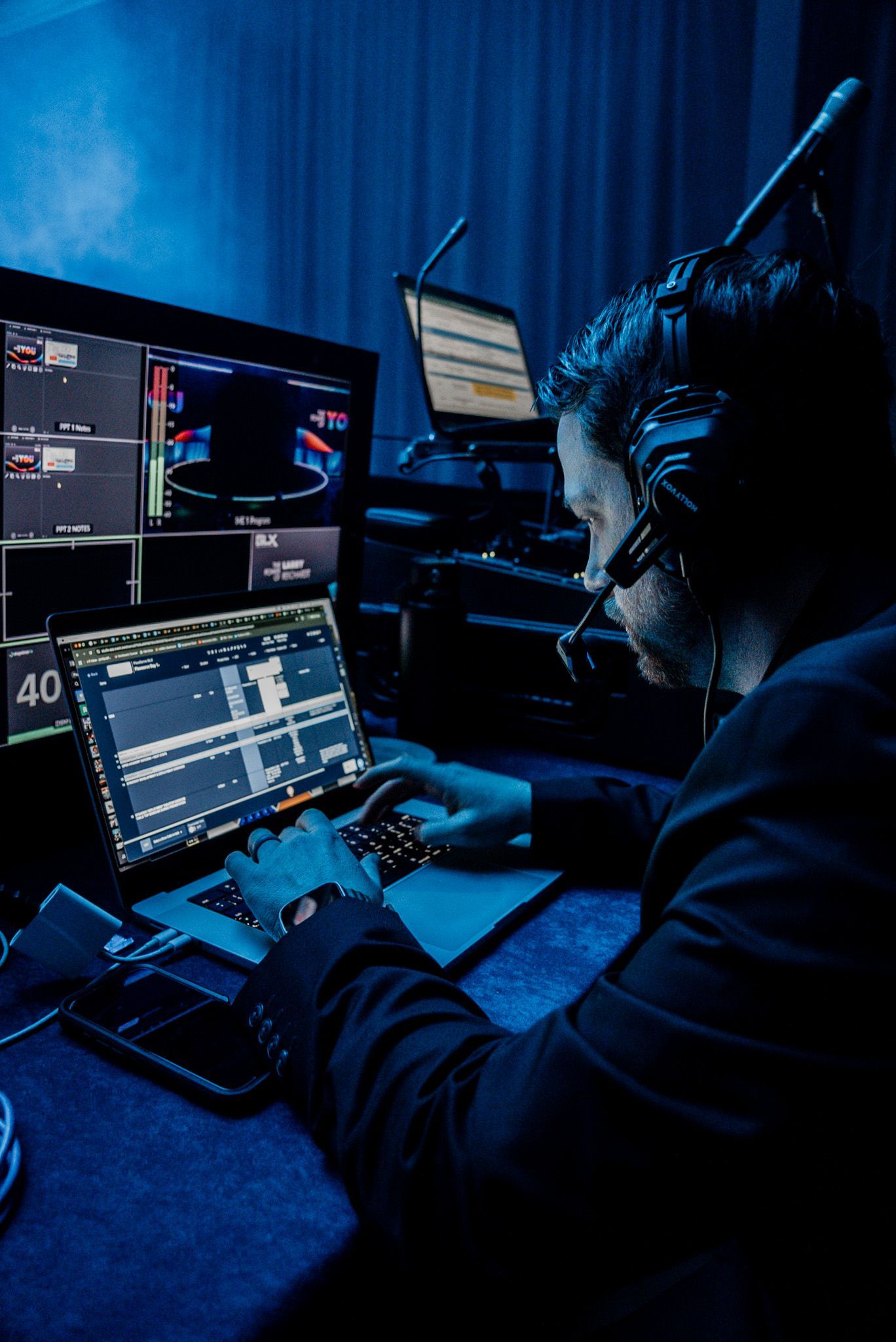 Person typing at a dual-monitor workstation in a dark blue-lit room, focused on a laptop and large screens.