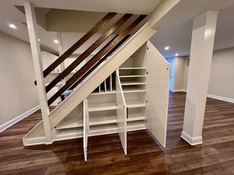Built-in storage under staircase with white cabinets, shelves, and brown railings in a room with hardwood floors.