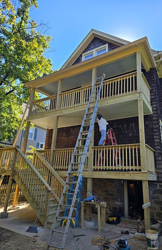 Two-story wooden deck addition to a house. Workers on ladders, tools below. Blue sky backdrop.