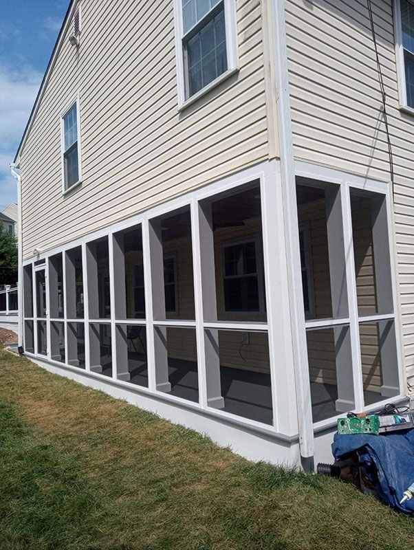 A screened-in porch with white trim and gray supports attached to a beige house, next to a green lawn.