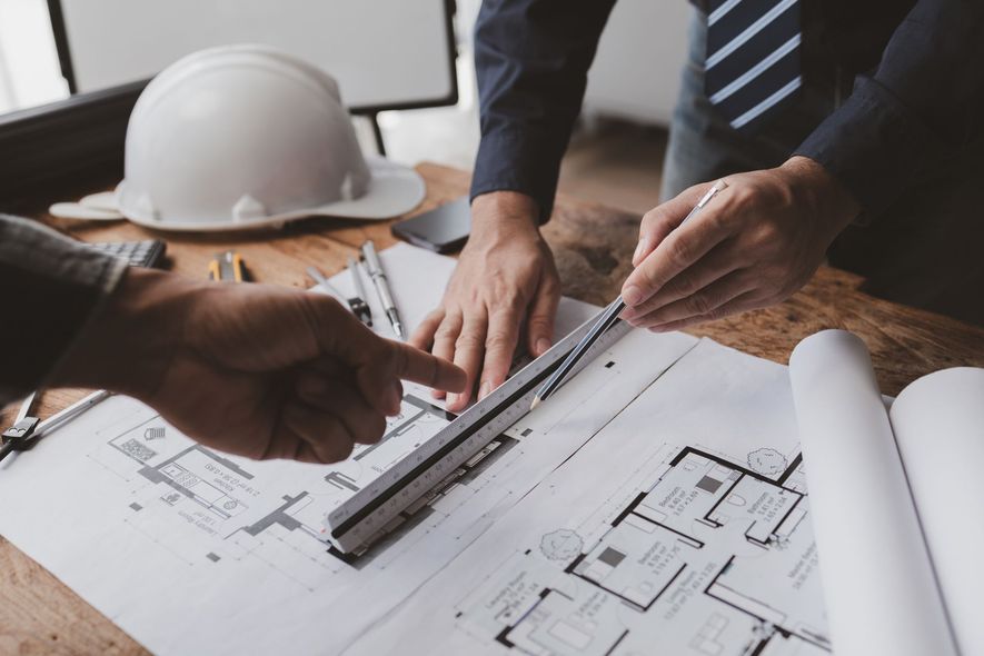 Two people reviewing architectural blueprints at a desk; one points, other uses a ruler.