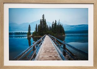 Wooden bridge leading to a small pine-covered island on a calm blue lake, framed by mountains