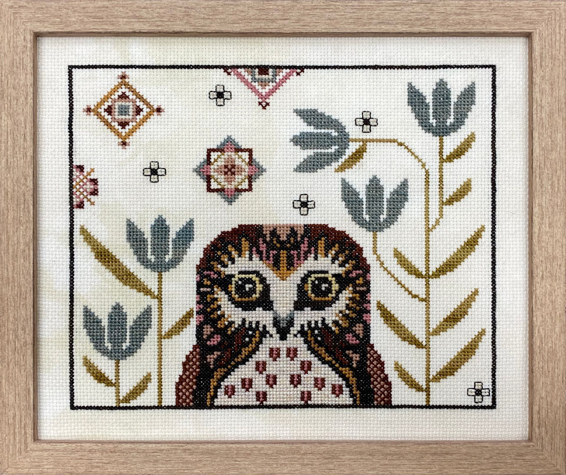 Handmade owl cross-stitch surrounded by colourful geometric shapes and botanical patterns in a natural wood frame.