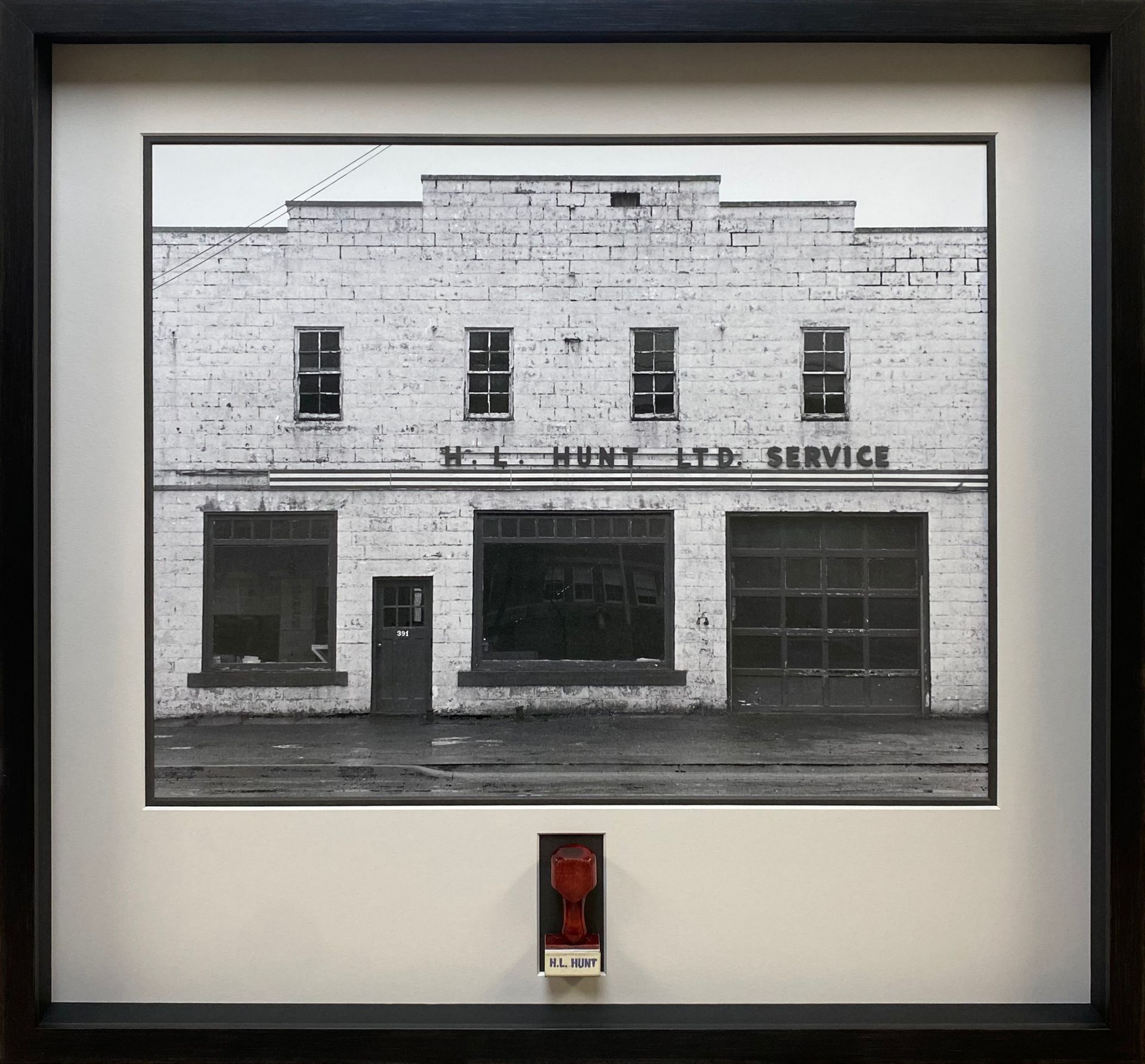 Black-and-white photo of an old storefront with a framed display and a small red plaque below