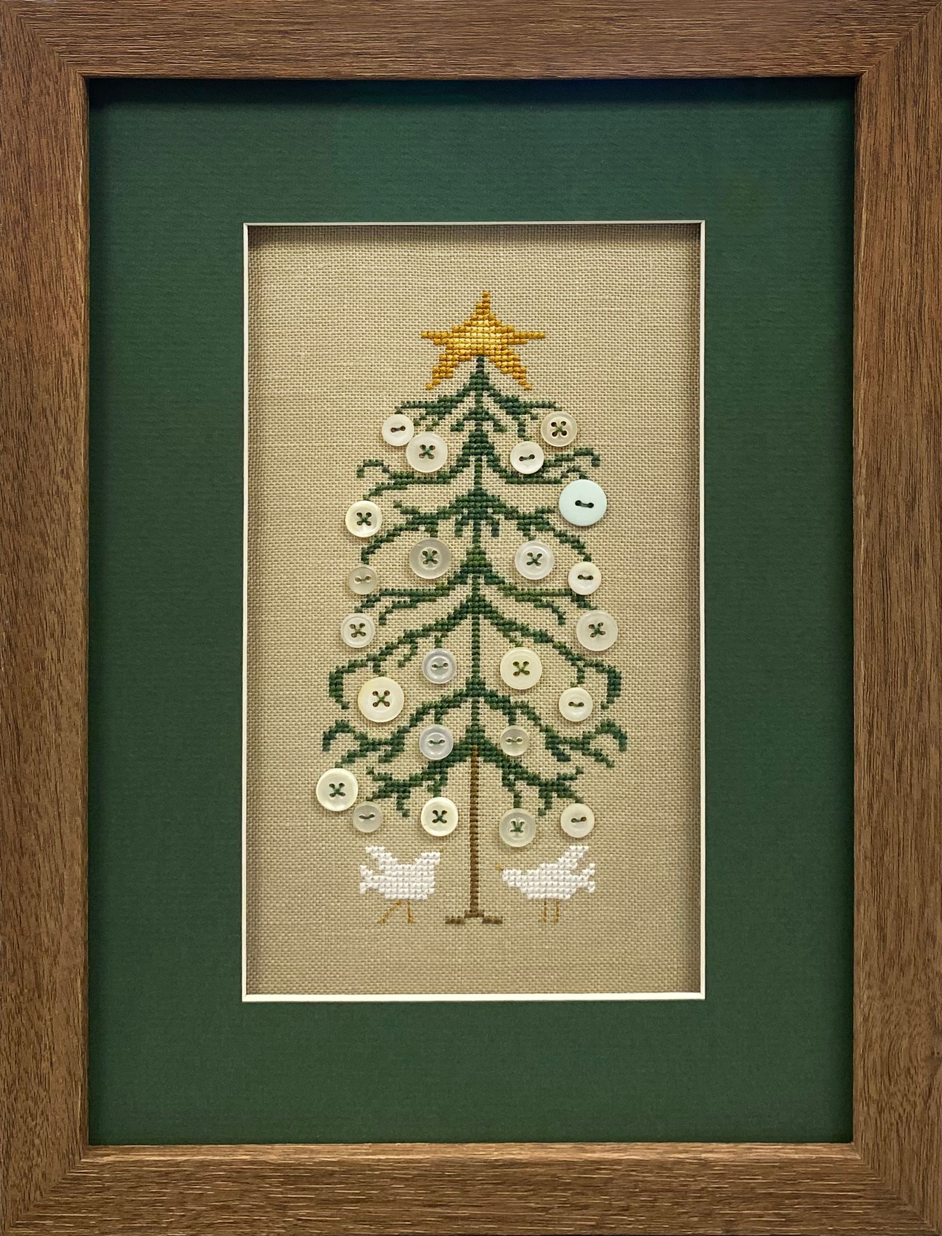 Handmade Christmas tree cross-stitch decorated with buttons, displayed with green matting in a wooden frame.
