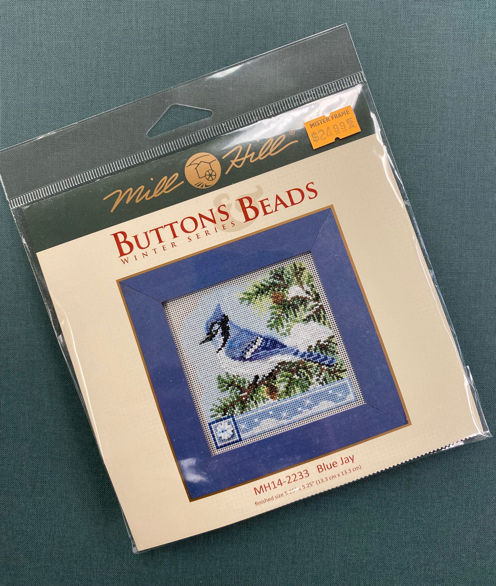 Needlework kit package with a blue bird design on a cream background, resting on green patterned fabric
