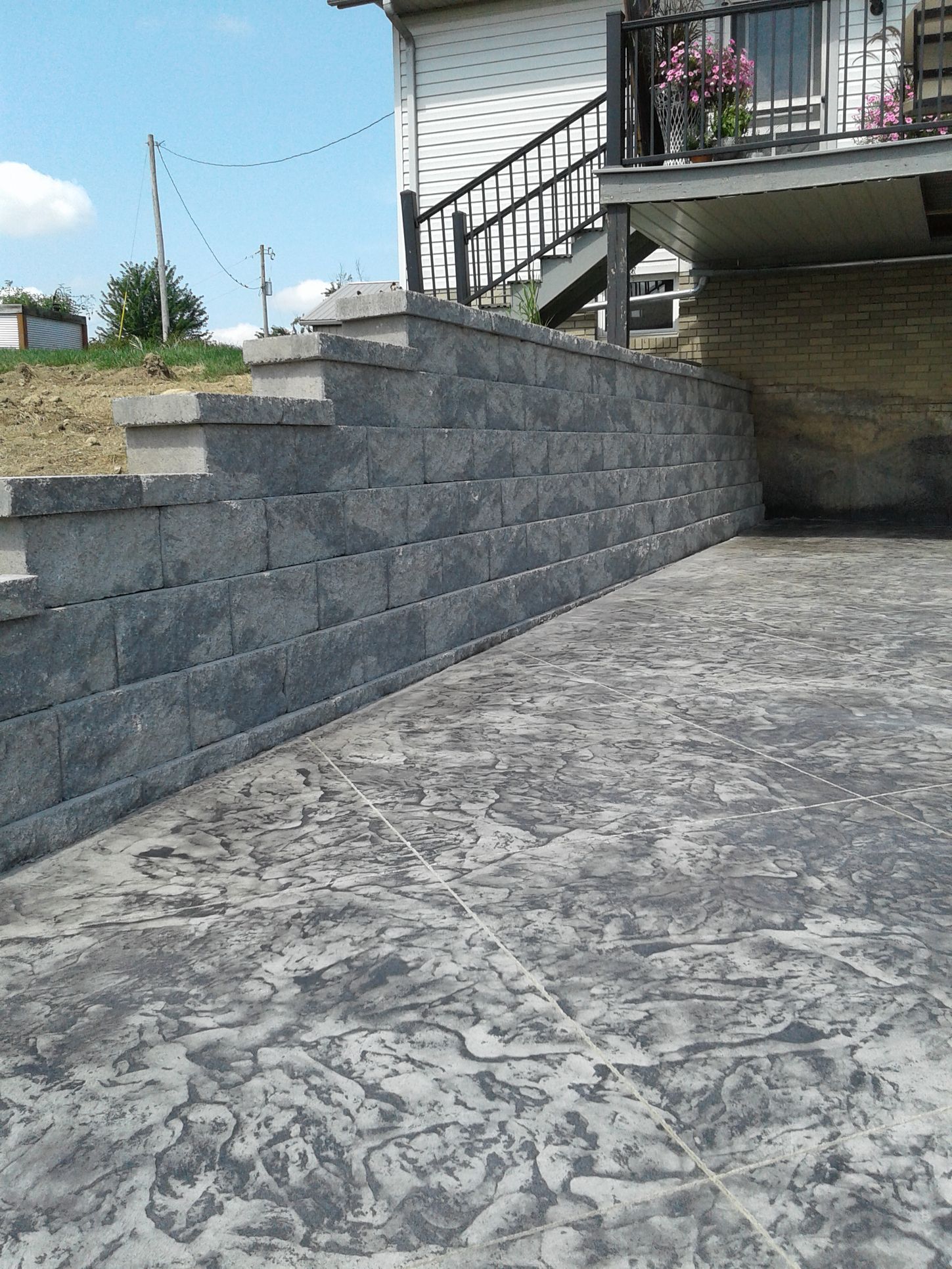 Gray retaining wall with steps, next to textured concrete patio. A house with a balcony is in the background.