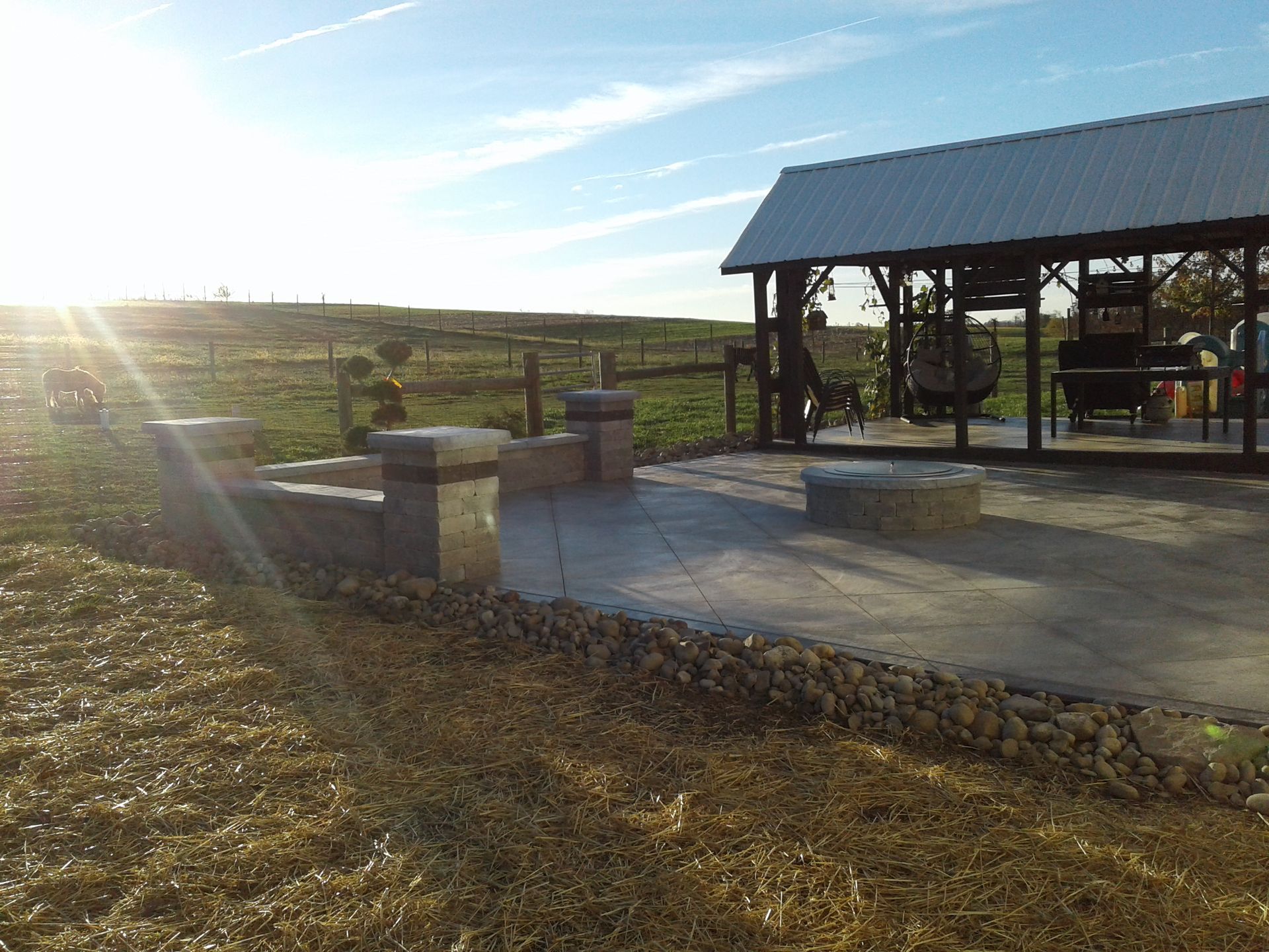 A sunlit outdoor concrete patio with a fire pit, and a metal-roofed shelter in a field.