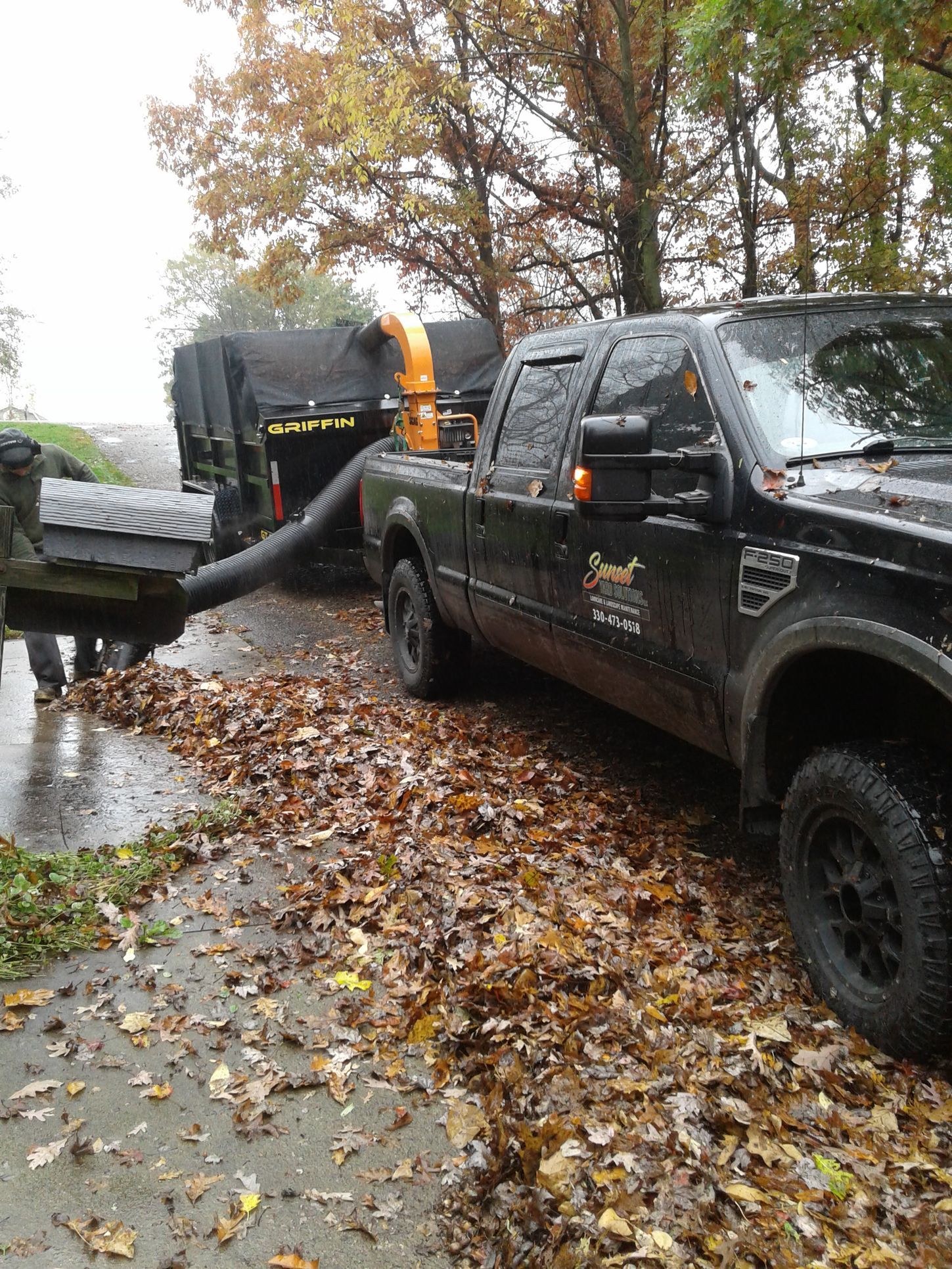 Truck vacuuming leaves on a wet road, leaves scattered around, trailer attached, autumn setting.