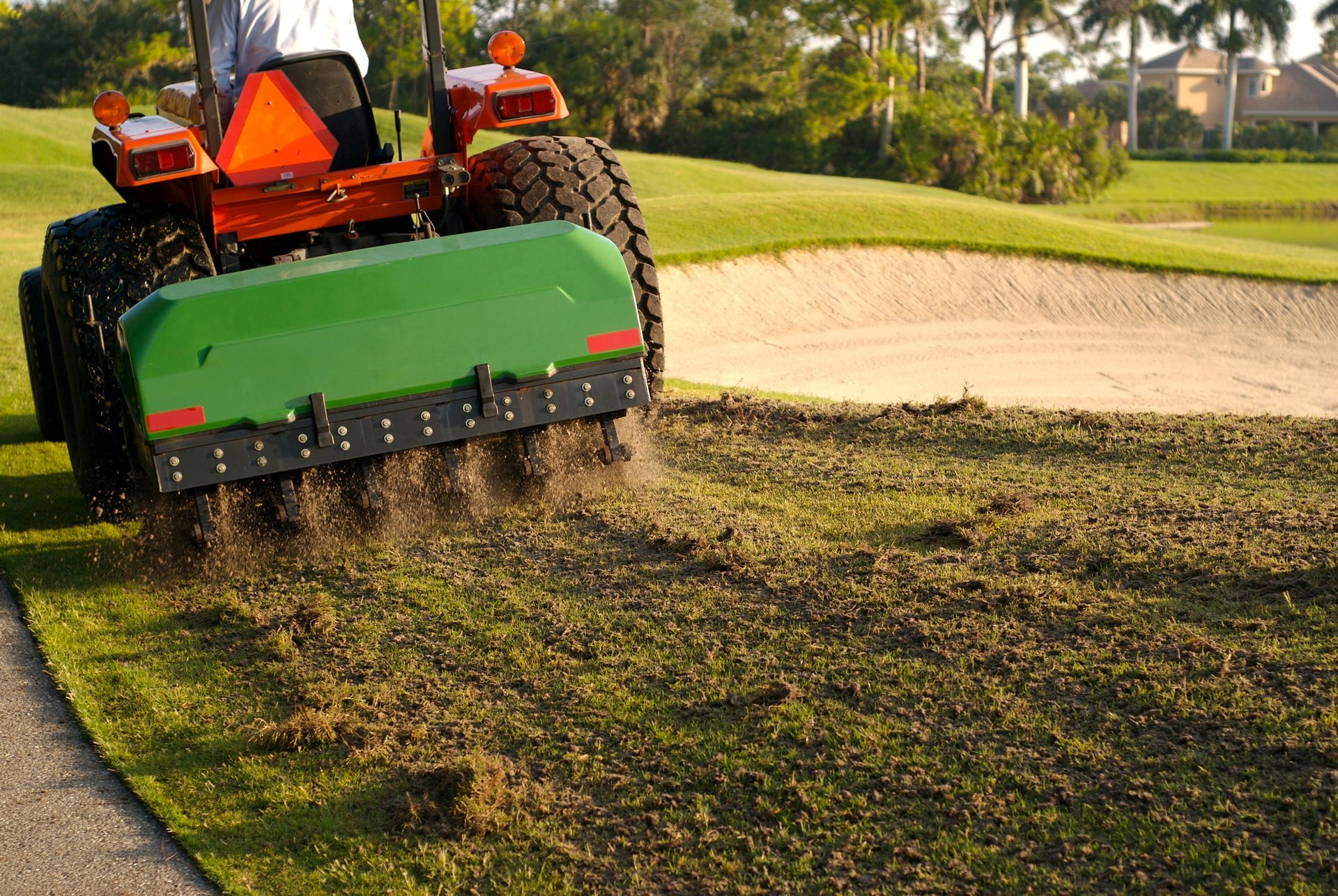 Tractor aerating green golf course. Green machine, brown soil, and sand bunker in the background.