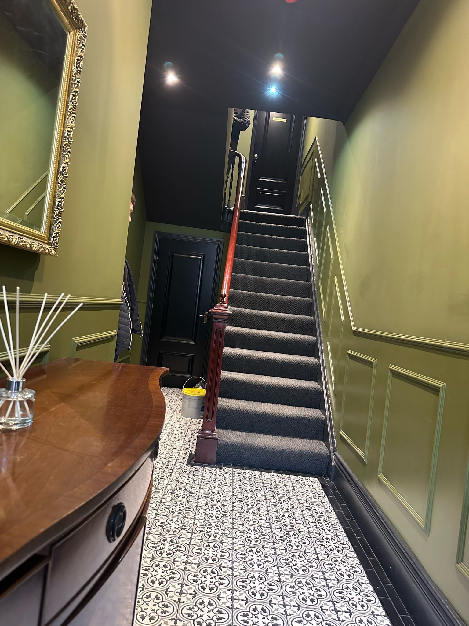 Narrow staircase in a dim hallway with black carpet, green walls, and patterned tile floor.