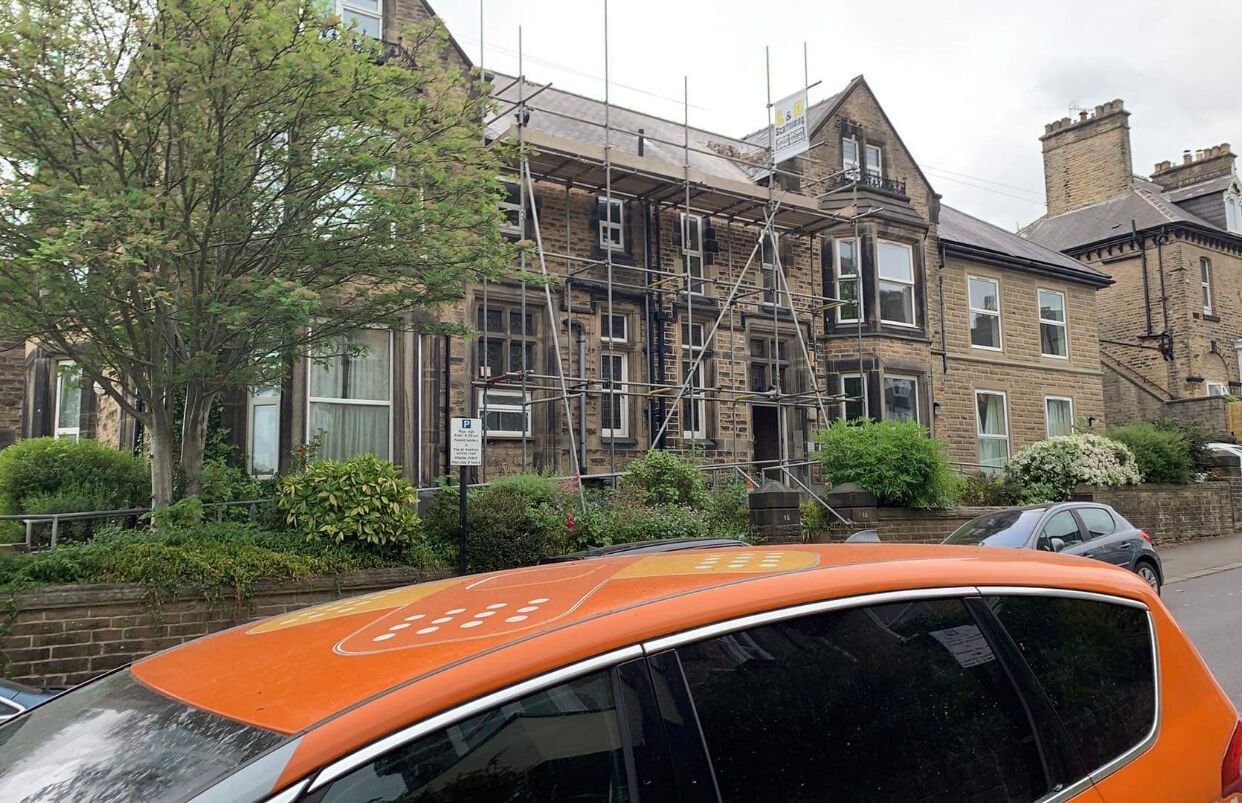 Row houses with scaffolding along a tree-lined street, partly blocked by an orange car in the foreground