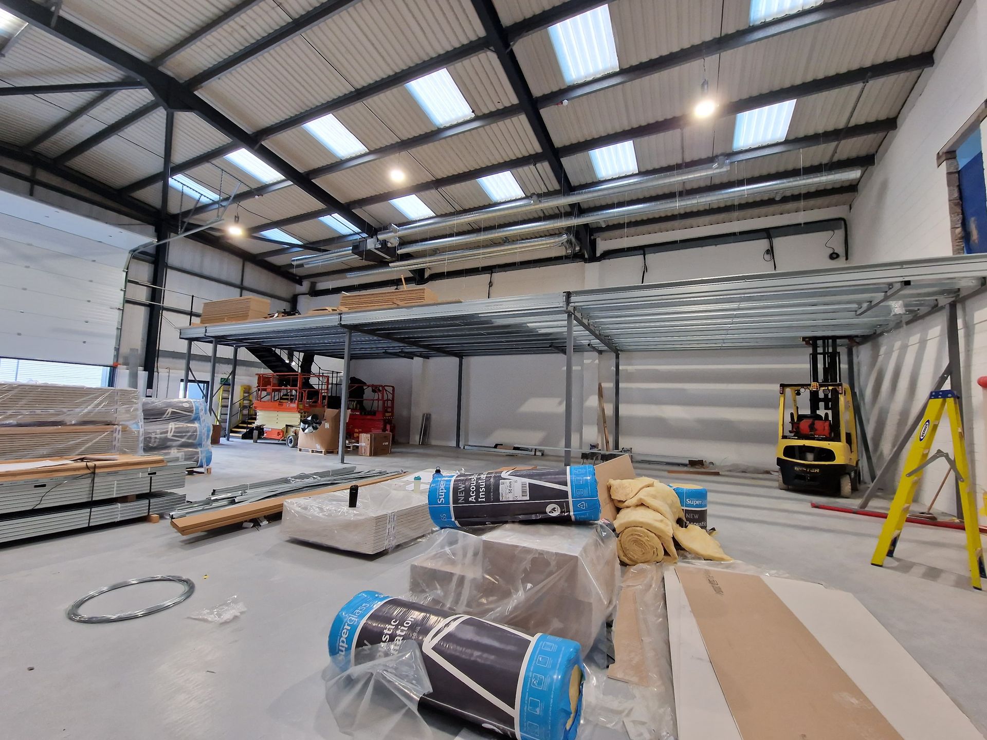 Large warehouse with a raised steel mezzanine under construction, pallets, tools, and a forklift in the foreground