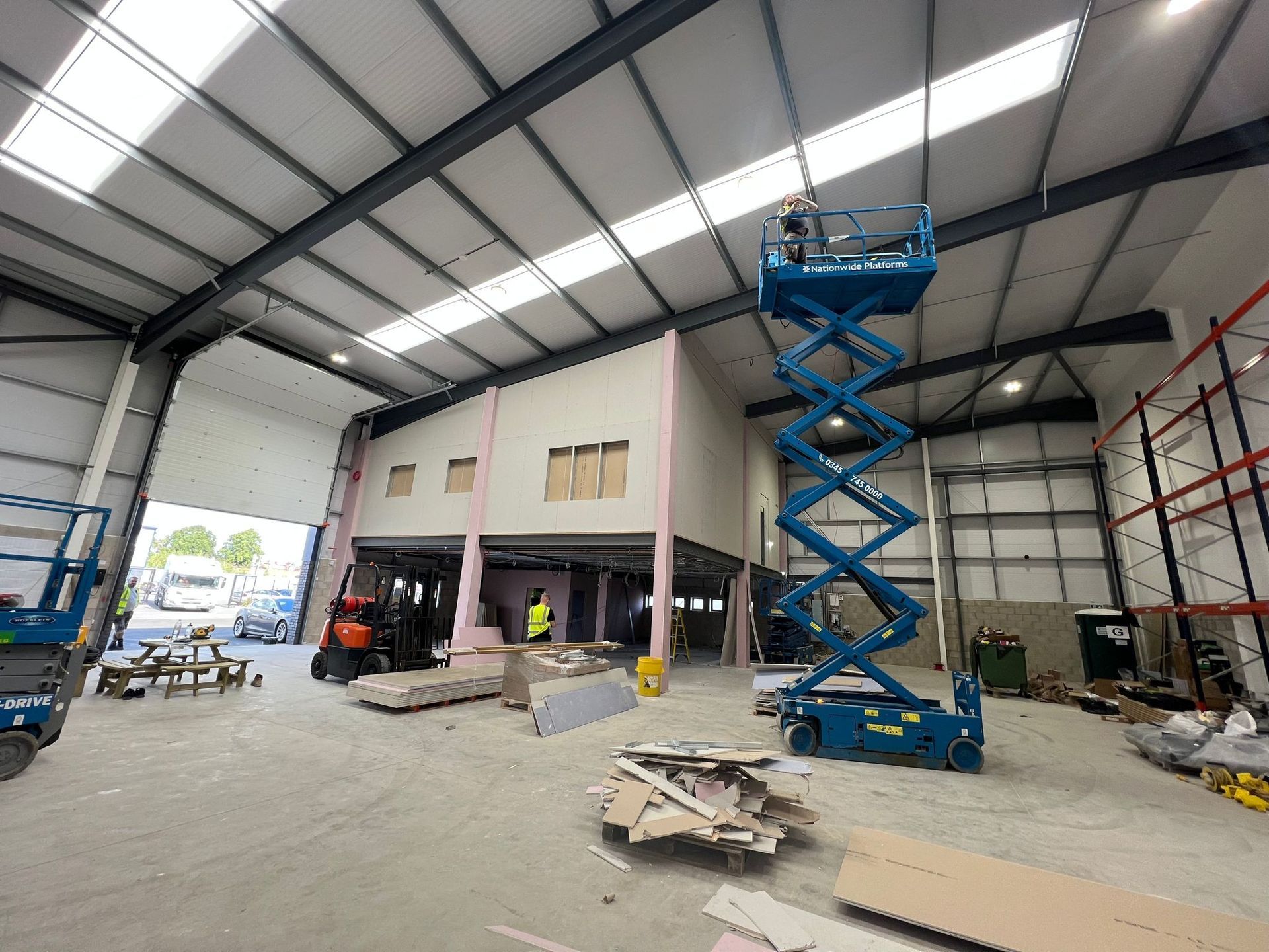 Spacious warehouse interior with a blue scissor lift, forklift, scaffolding, and scattered building materials.