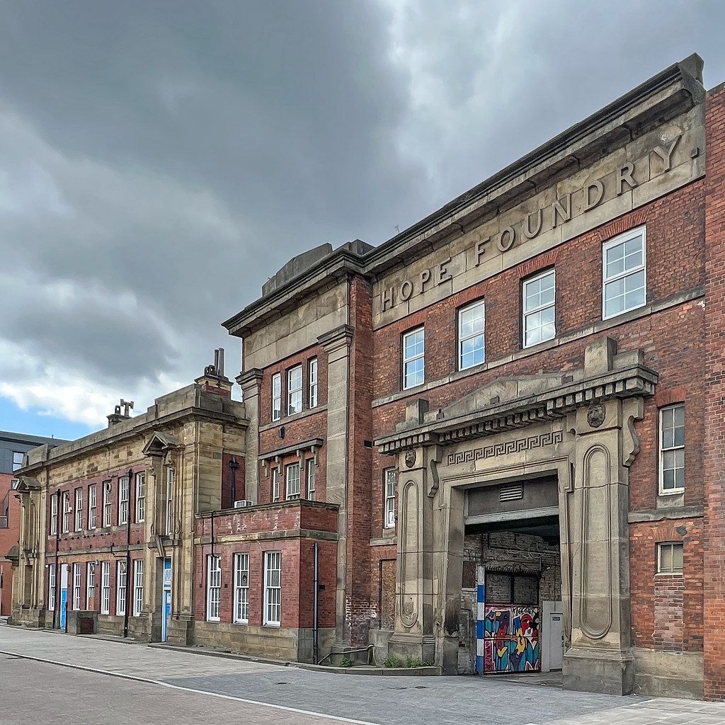 Dilapidated red-brick industrial building with boarded windows and a large arched entrance under a cloudy sky