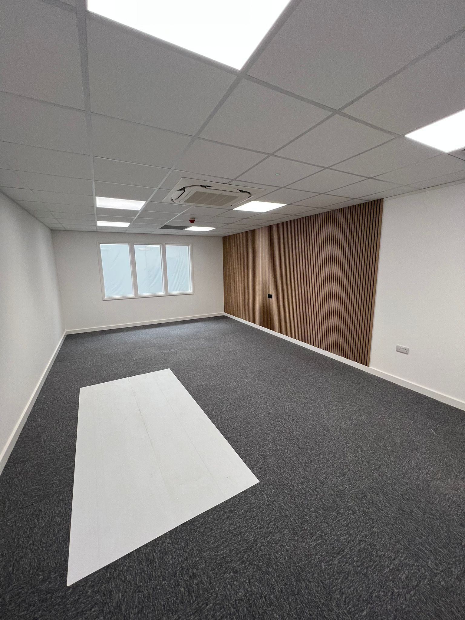 Empty office room with gray carpet, white ceiling tiles, windows, and a wood-patterned accent wall.
