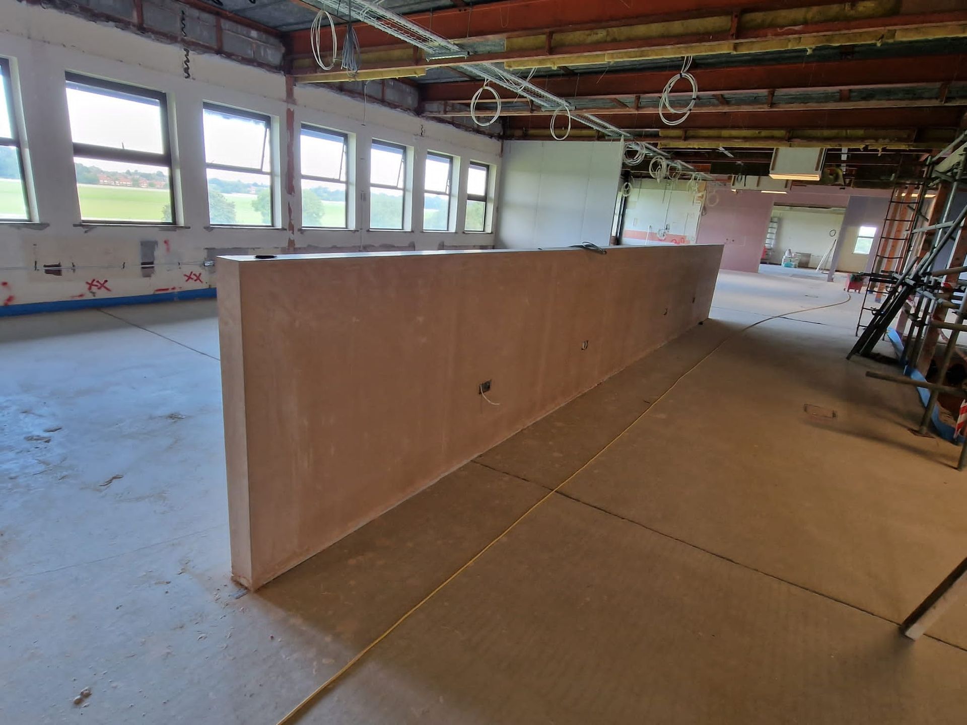 A long, plastered partition wall stands in a large, unfinished commercial space with exposed ceiling beams and windows.