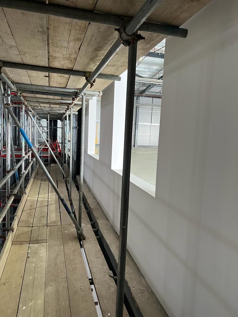 Scaffolding in a narrow indoor corridor beside white walls and exposed ceiling beams.
