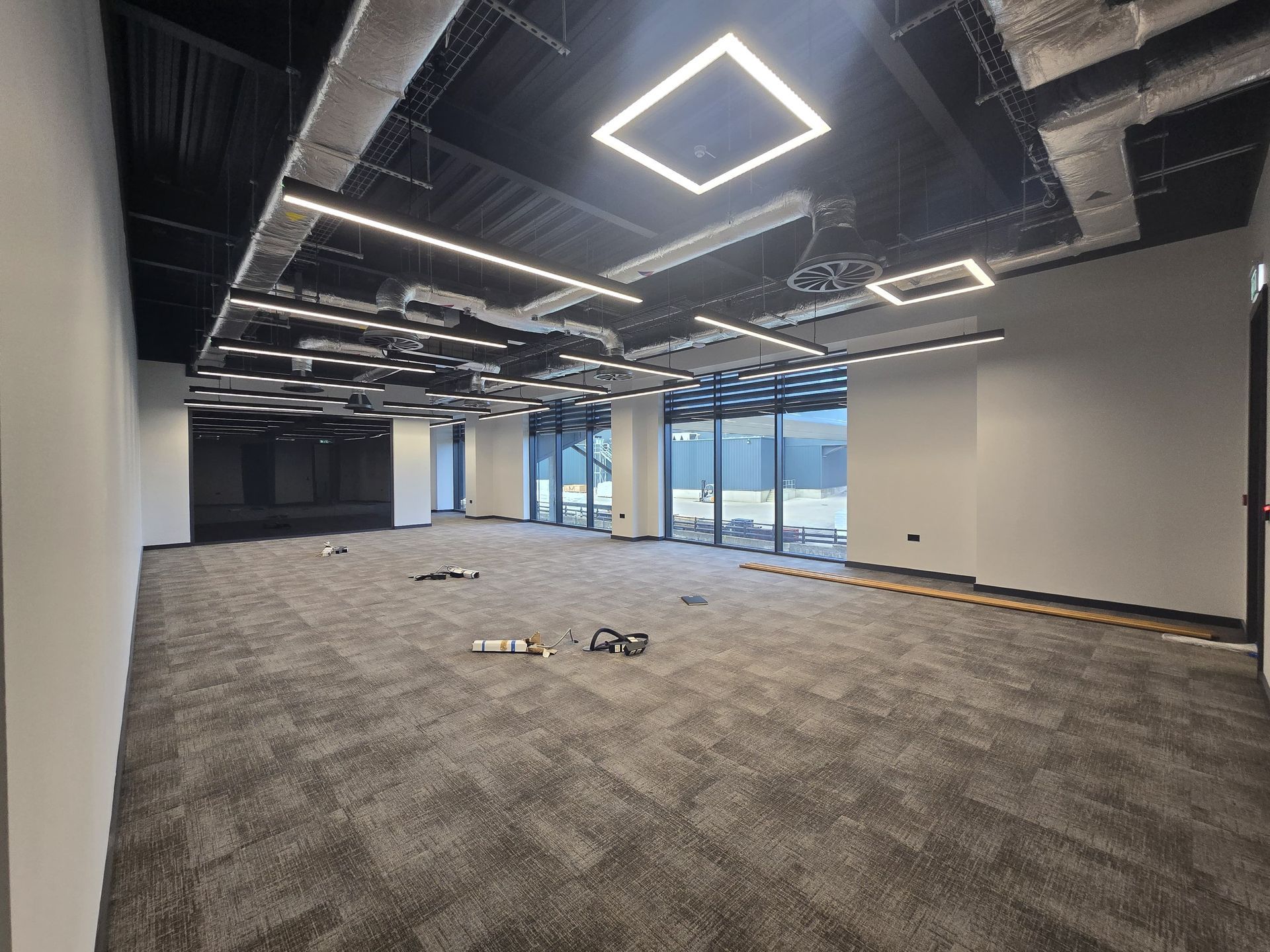 Empty modern office space with carpeted floor, exposed ceiling lights, and large windows.