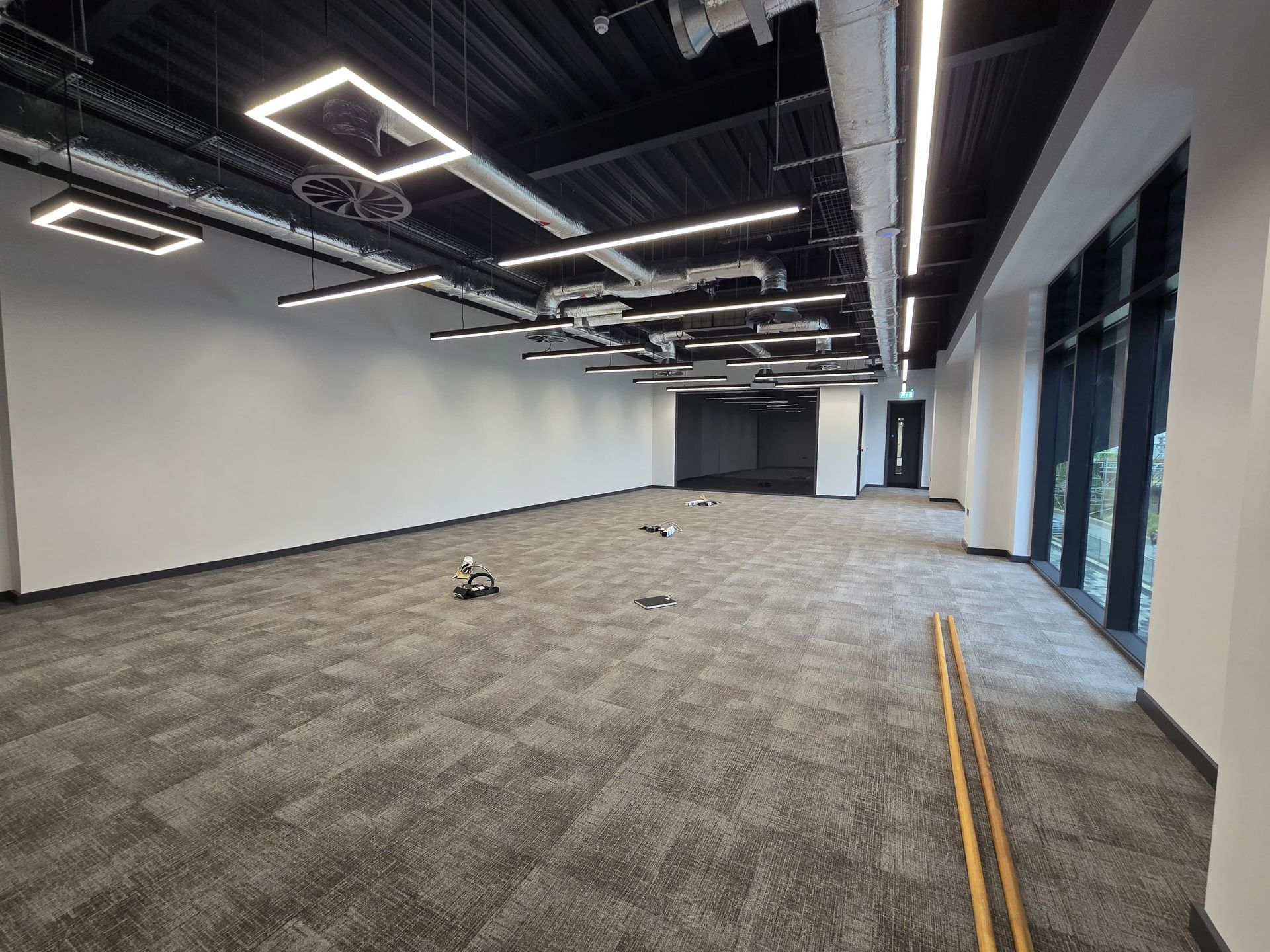 Empty modern office space with gray carpet, white walls, glass doors, and overhead strip lighting.