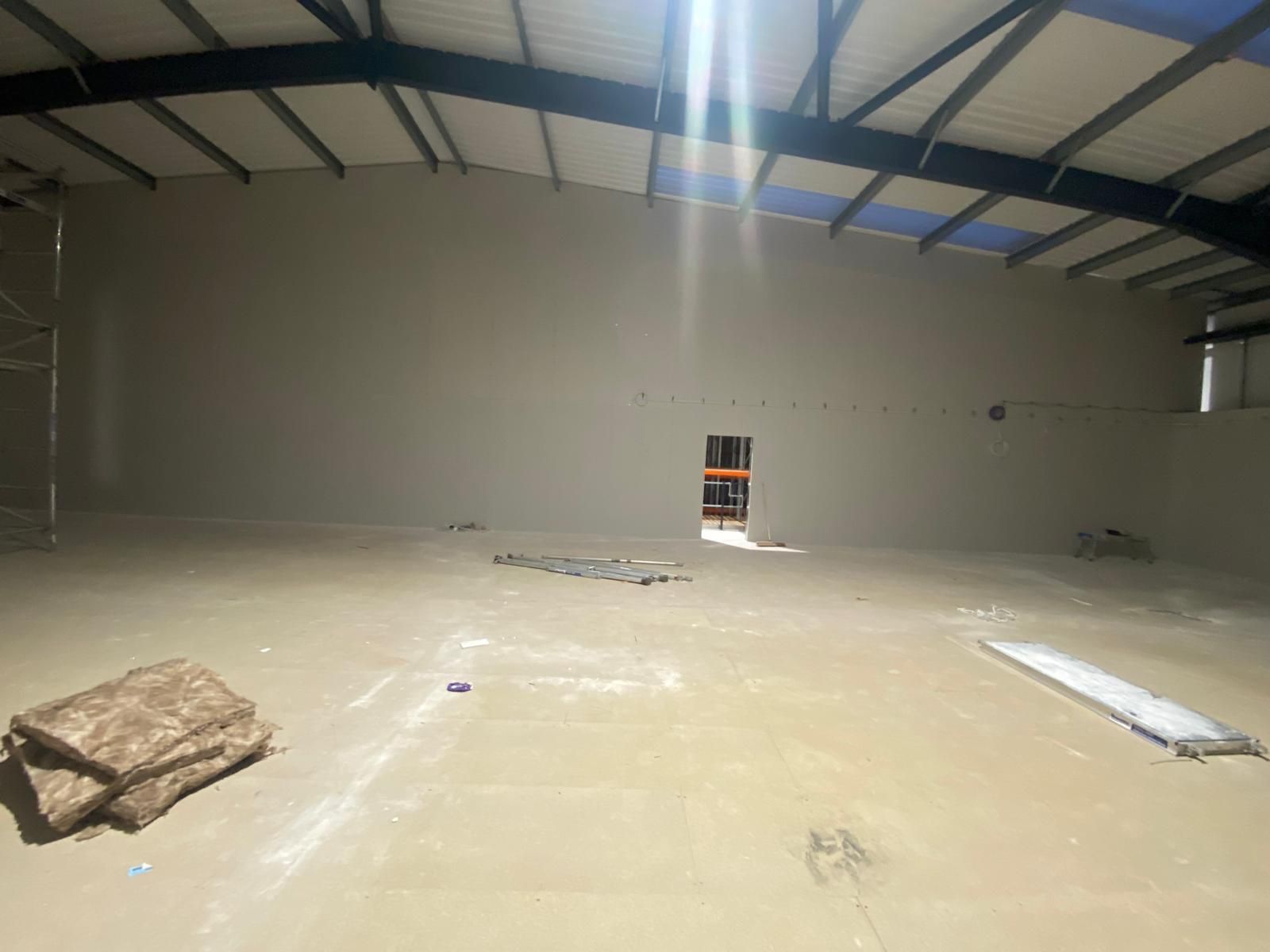Large empty warehouse with a concrete floor, exposed roof beams, and a small doorway in the far wall.