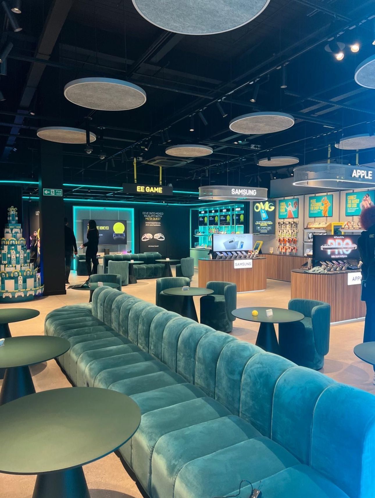 Modern lounge with teal couches, round ceiling lights, and glowing arcade-style game machines in the background