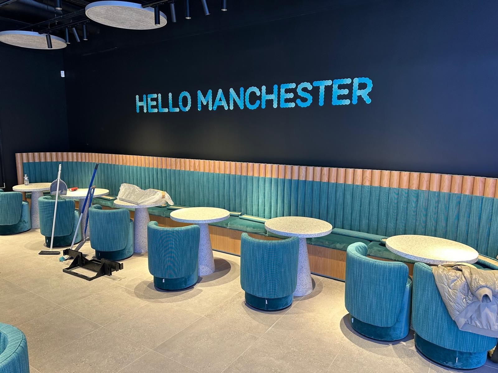 Blue booth seating and round tables in a modern lounge with “HELLO MANCHESTER” on the wall.