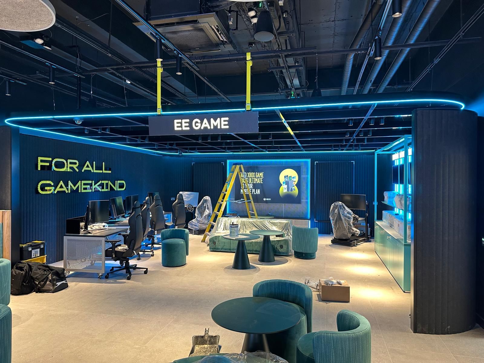 Futuristic gaming lounge with neon blue lighting, desks, chairs, and large wall text reading “BE GAME”