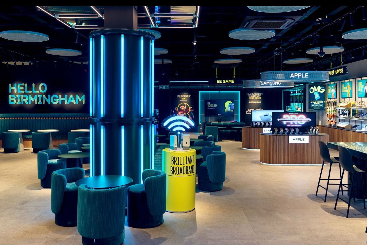 Neon-lit airport lounge with teal chairs, a yellow charging station, and “HELLO BIRMINGHAM” on the wall
