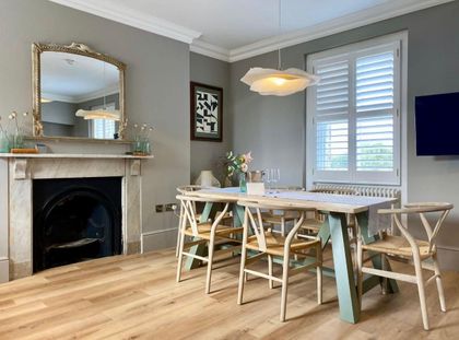 Bright dining room with a rustic table, chairs, fireplace, mirror, and window shutters