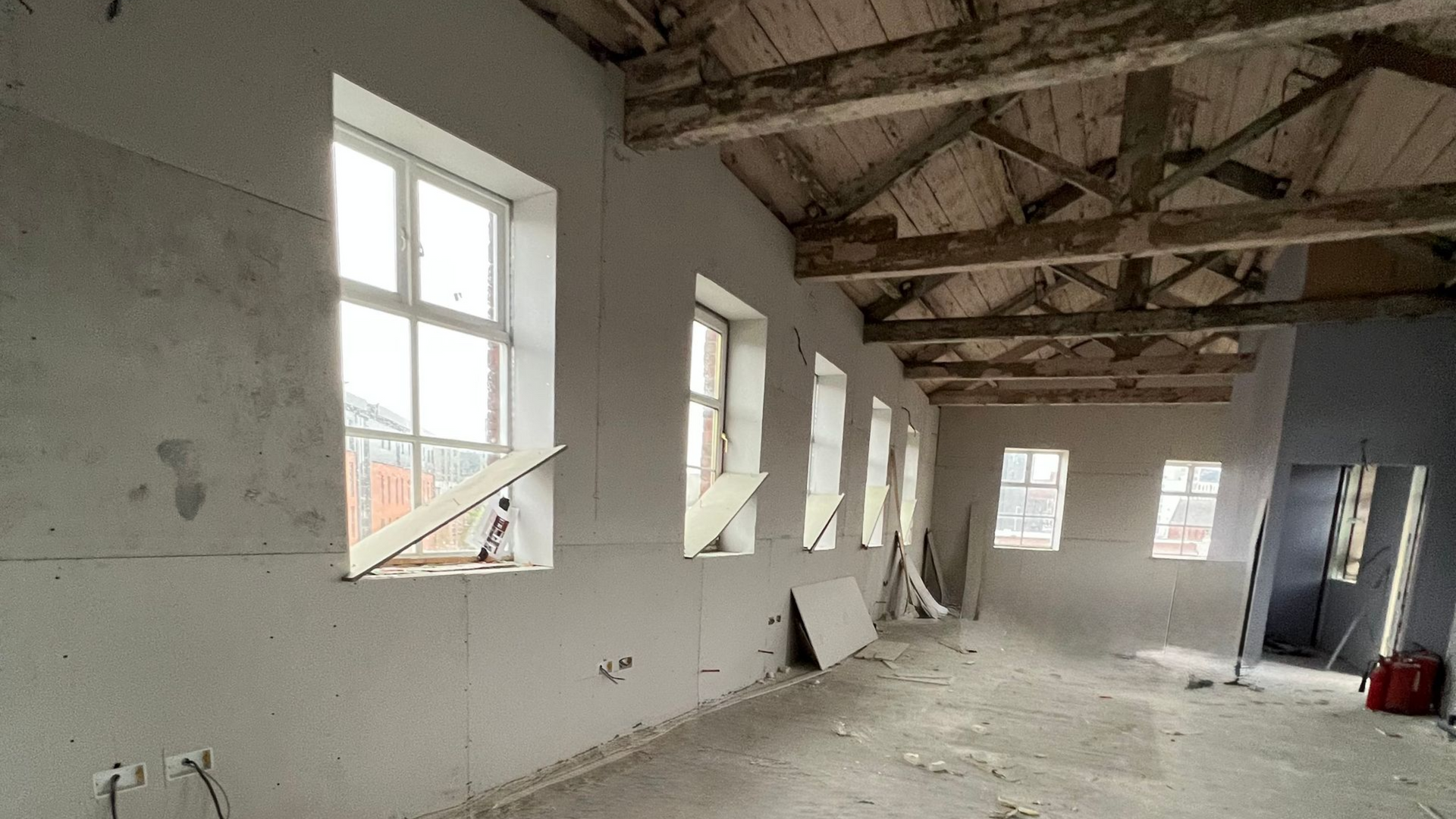 A room under construction with gray drywall walls, multiple windows, and exposed wooden ceiling beams.