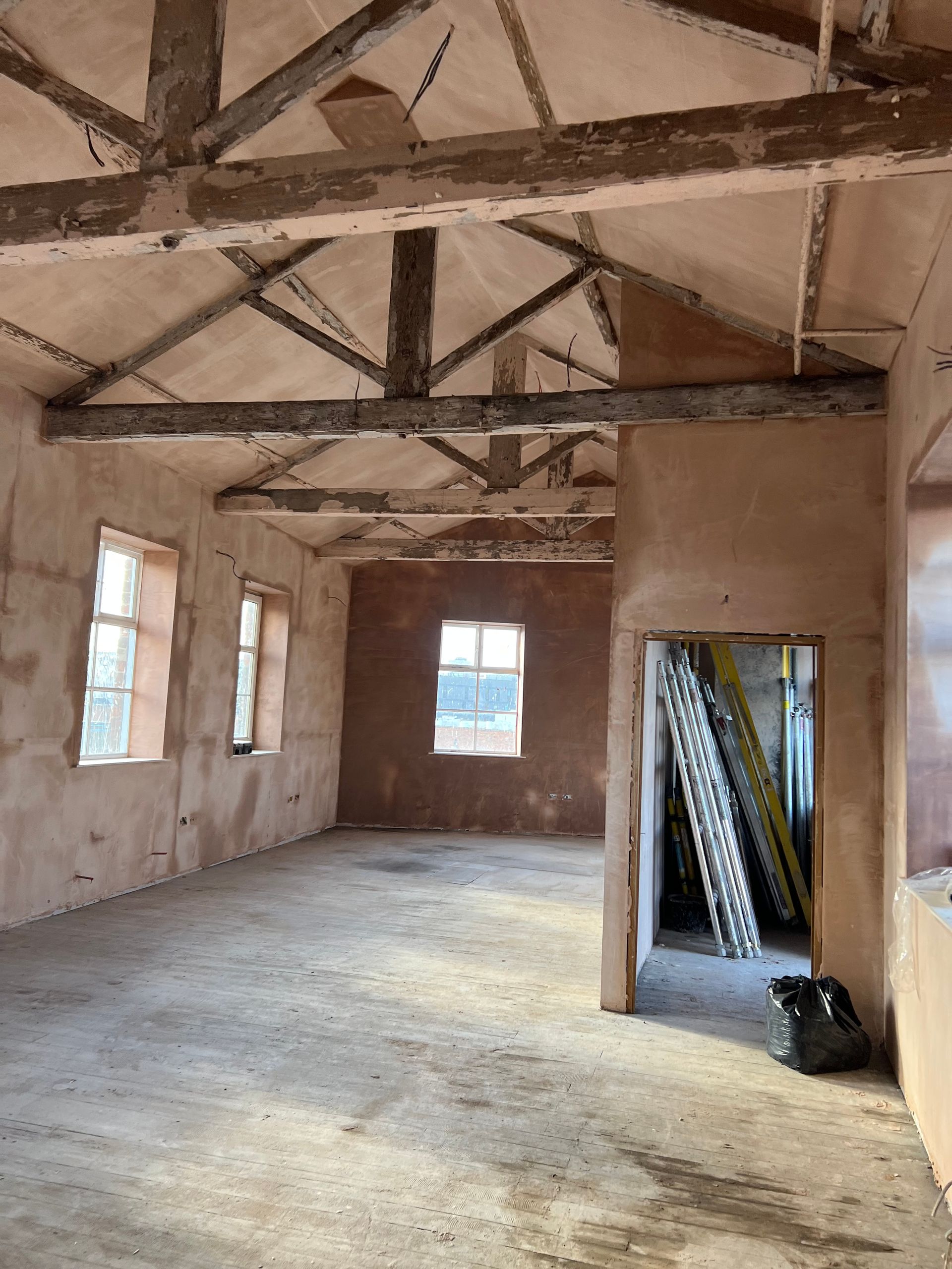 Empty unfinished room with exposed beams, plaster walls, windows, and ladder leaning near an open doorway.