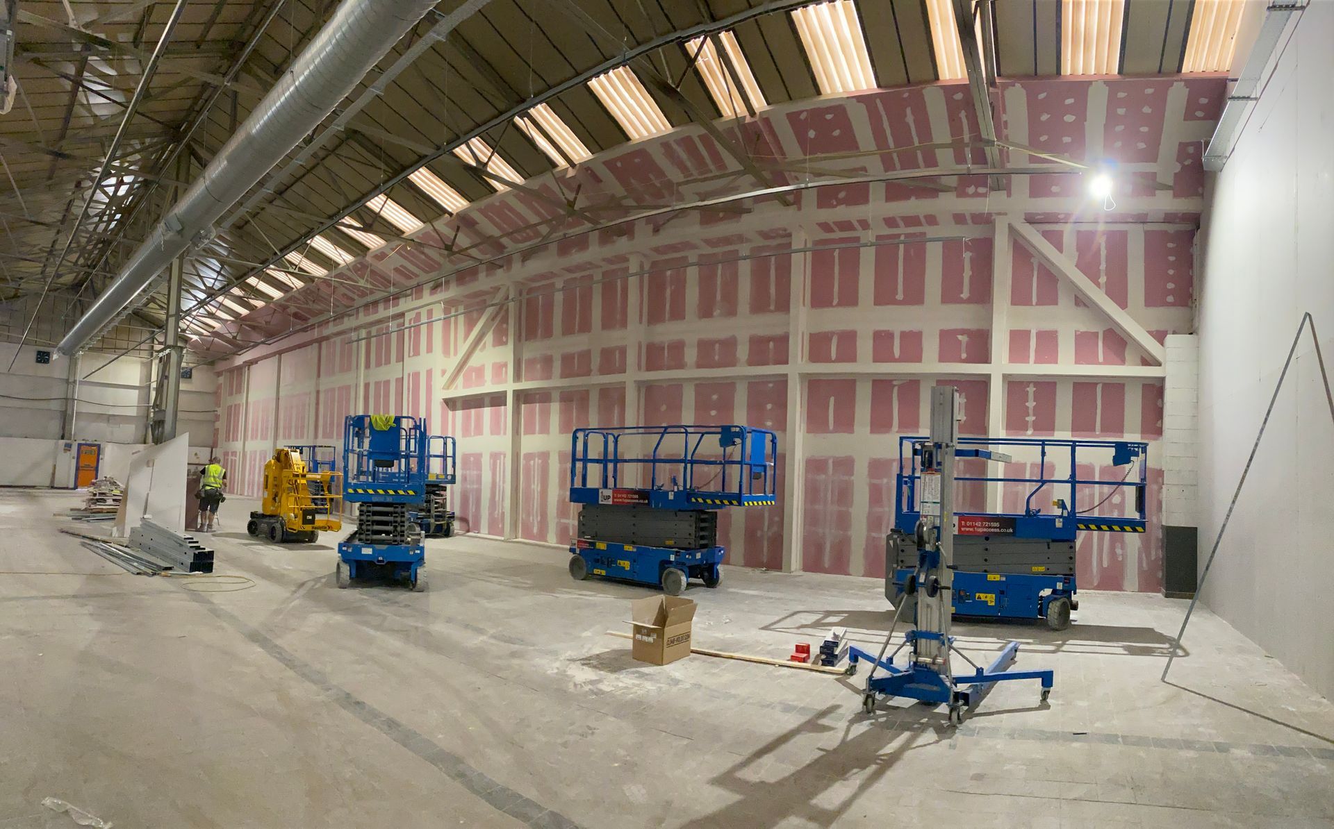Construction site interior with scaffolding lifts and exposed pink insulated wall under a curved roof