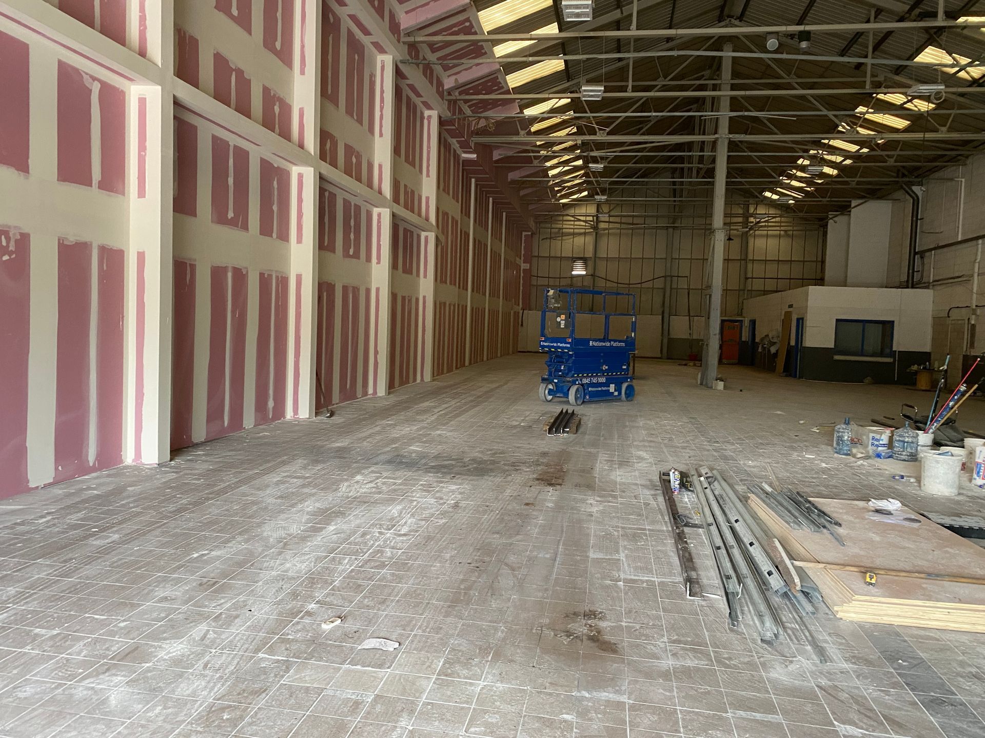 Empty warehouse interior with unfinished drywall, construction debris, and a blue scissor lift in the center