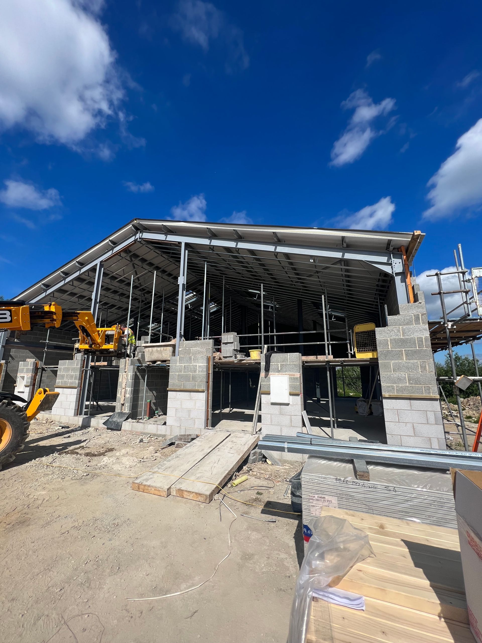Construction site with steel-framed building under blue sky, fronted by concrete blocks and machinery