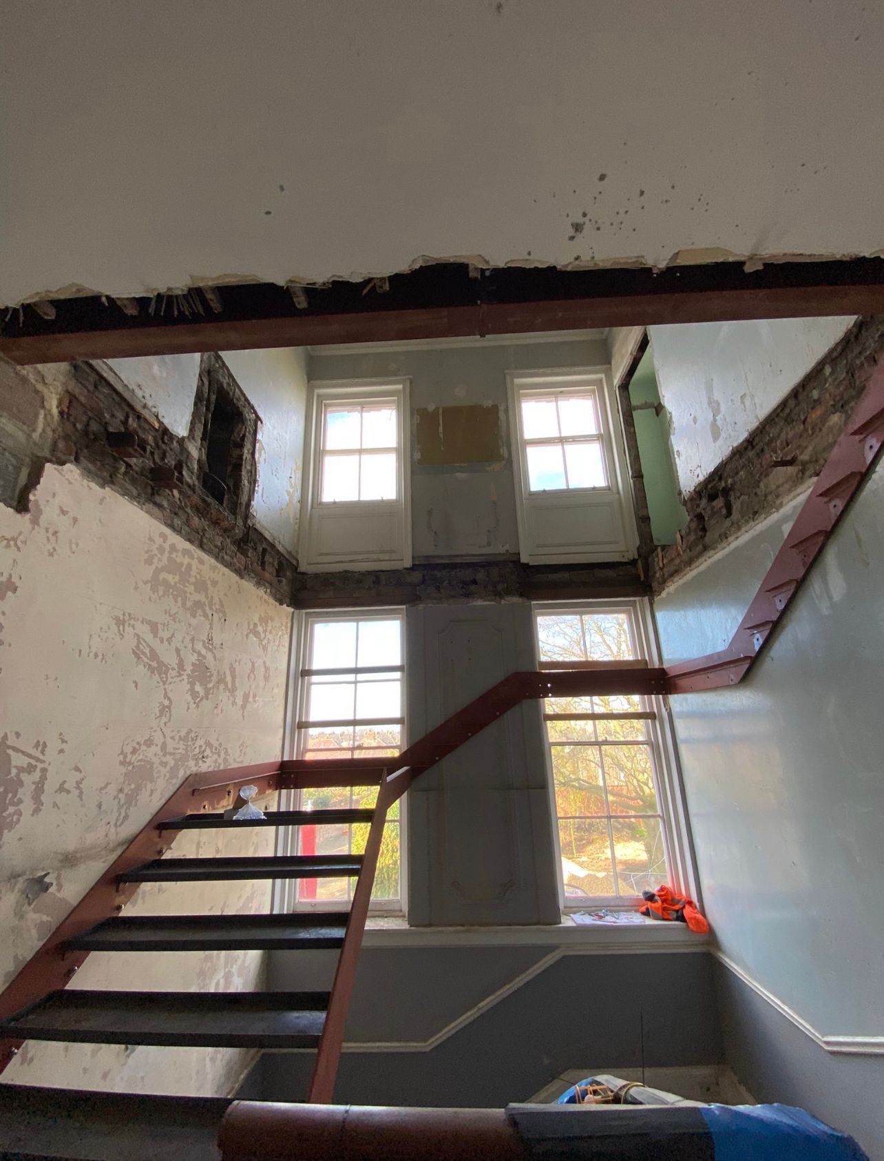 Damaged indoor staircase in a narrow stairwell with peeling walls and exposed beams, lit by two windows.