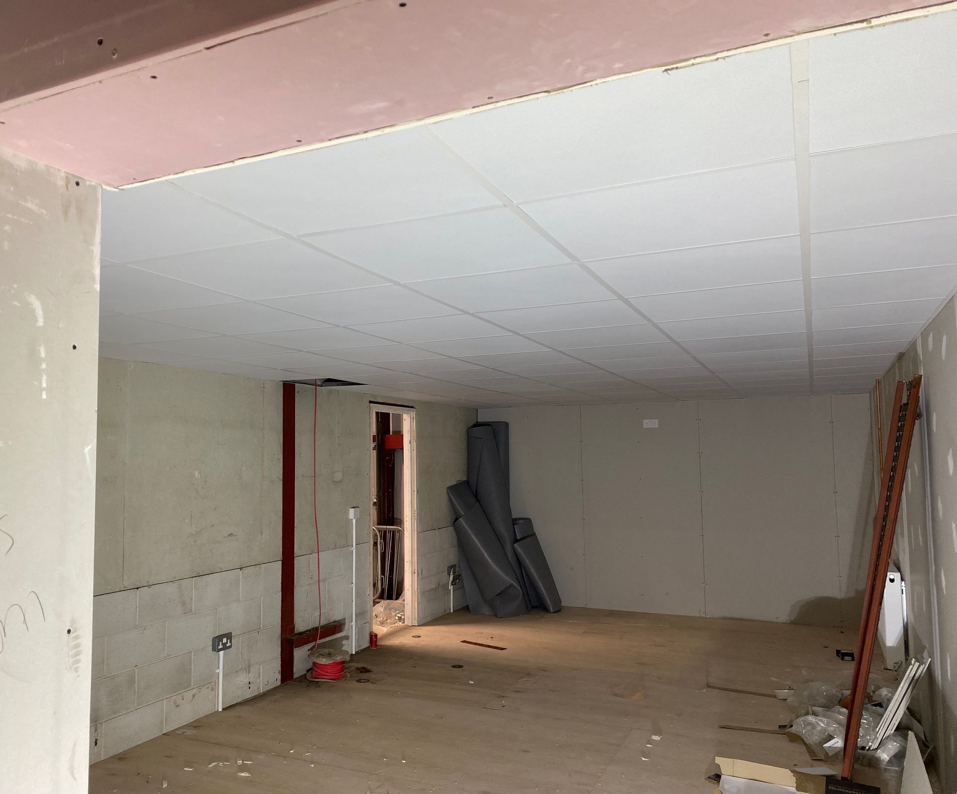 Empty unfinished room with white ceiling tiles, bare walls, exposed doorway, and construction materials.