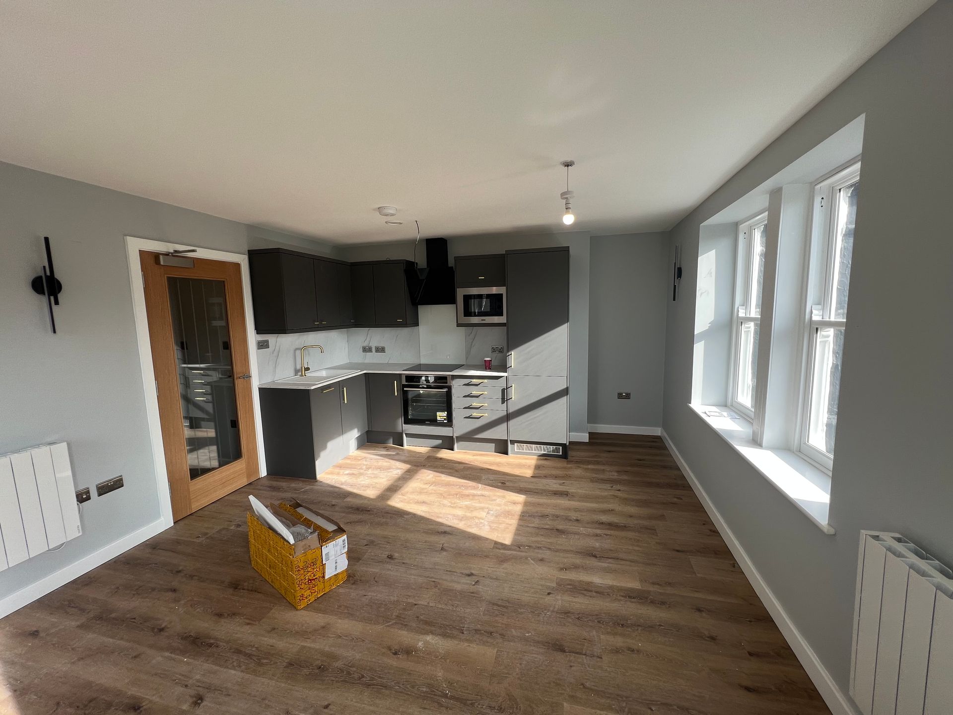 Empty modern kitchen and living area with white walls, gray cabinets, and large windows.