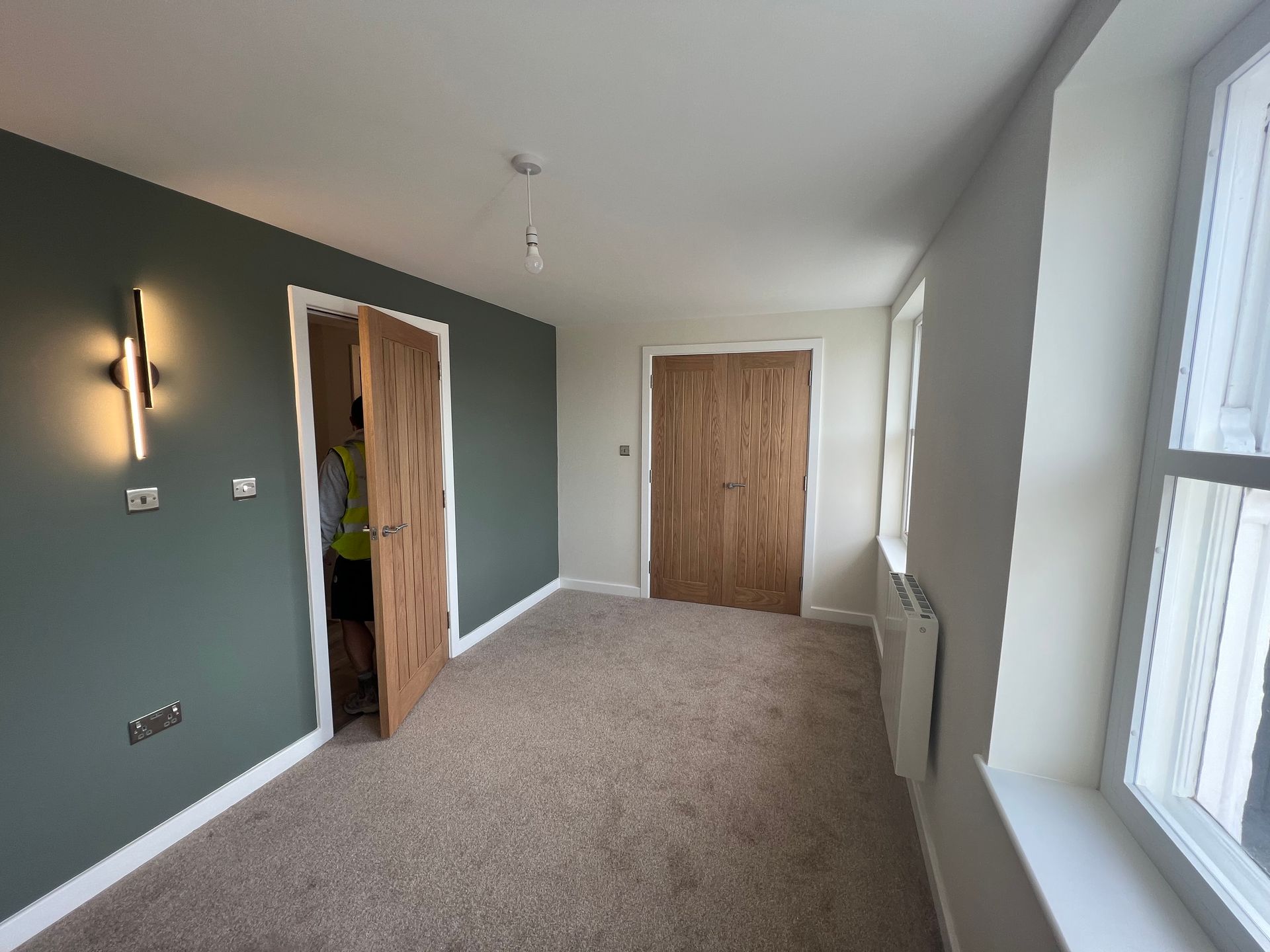 Empty carpeted room with green accent wall, two wooden doors, and large windows on the right.
