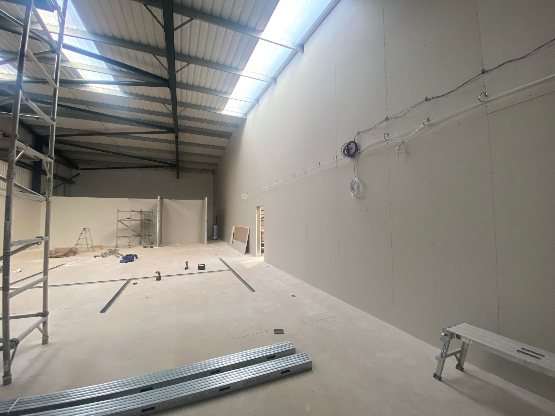 Large unfinished warehouse interior with white walls, exposed roof, scaffolding, and construction materials on the floor