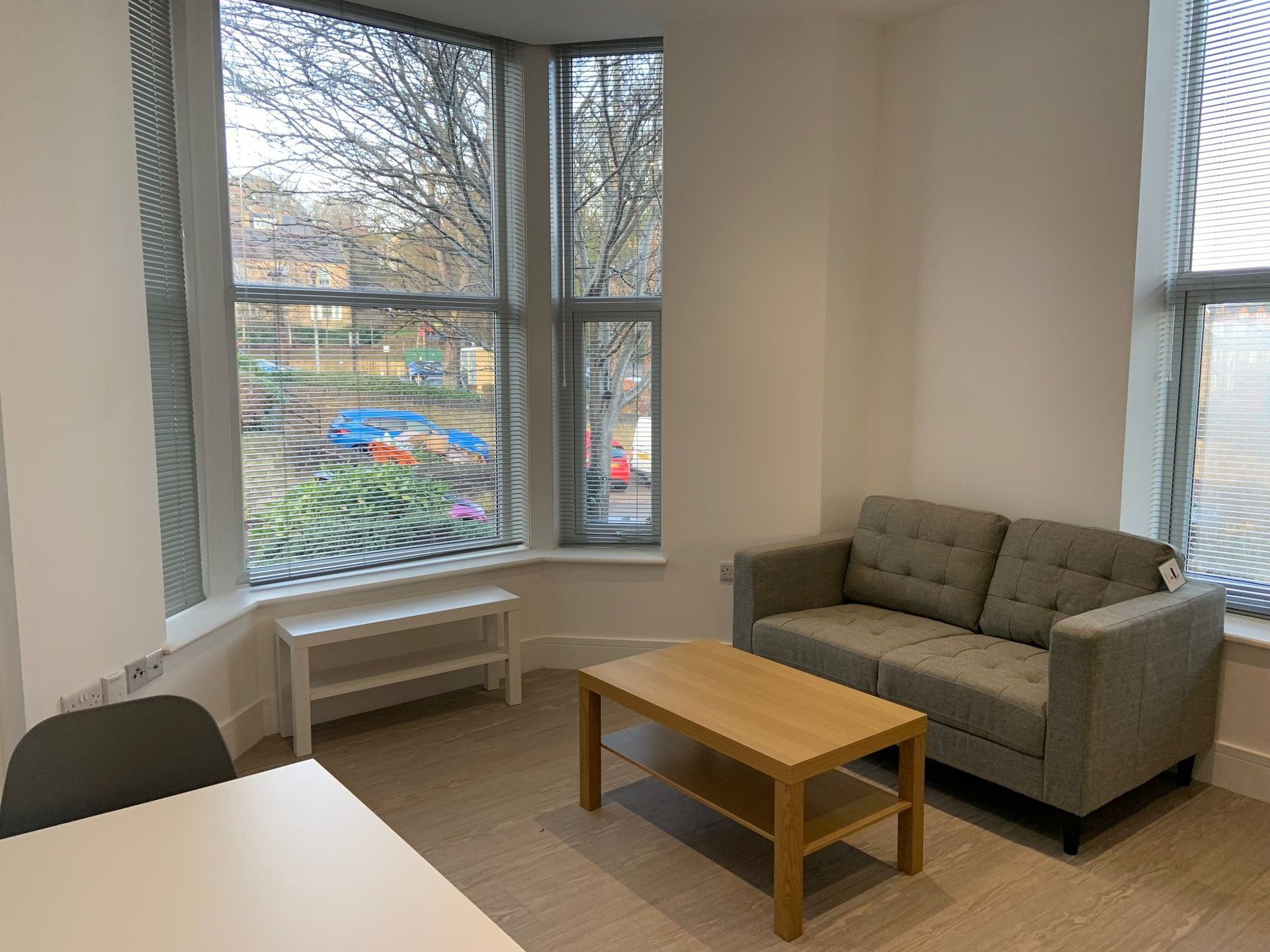 Small office lounge with beige sofa, wooden coffee table, white bench, and large windows overlooking trees