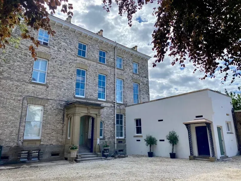 Stone building with blue windows beside a white annex, viewed from a courtyard under leafy trees.