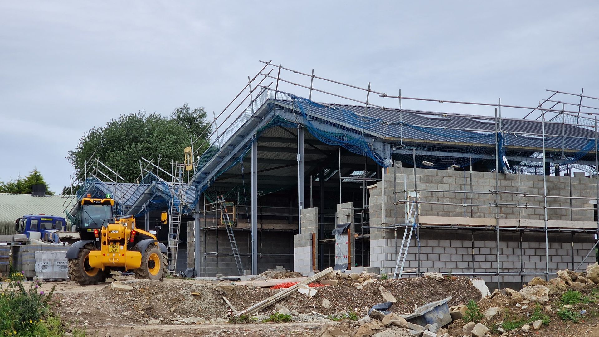 Construction site with a yellow excavator beside a partially built concrete structure and blue safety netting.