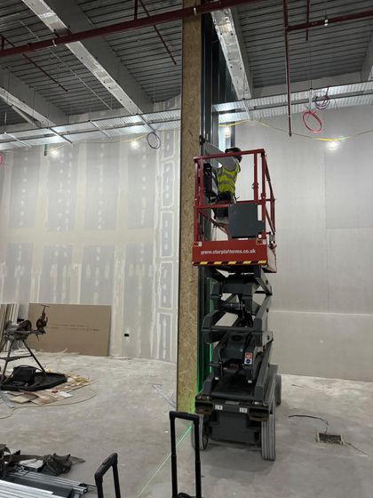 Worker on a red scissor lift inside an unfinished room with drywall and exposed ceiling beams.
