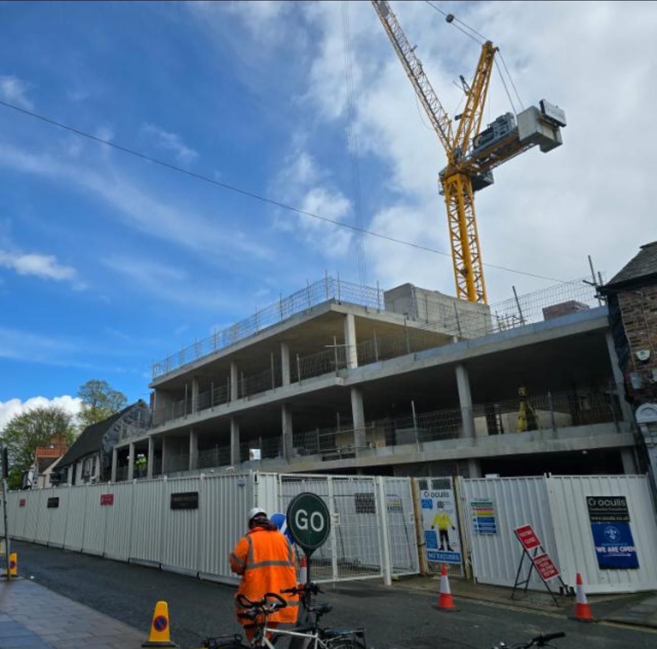 Construction site with crane, unfinished building, and worker in orange vest by a fence under a blue sky