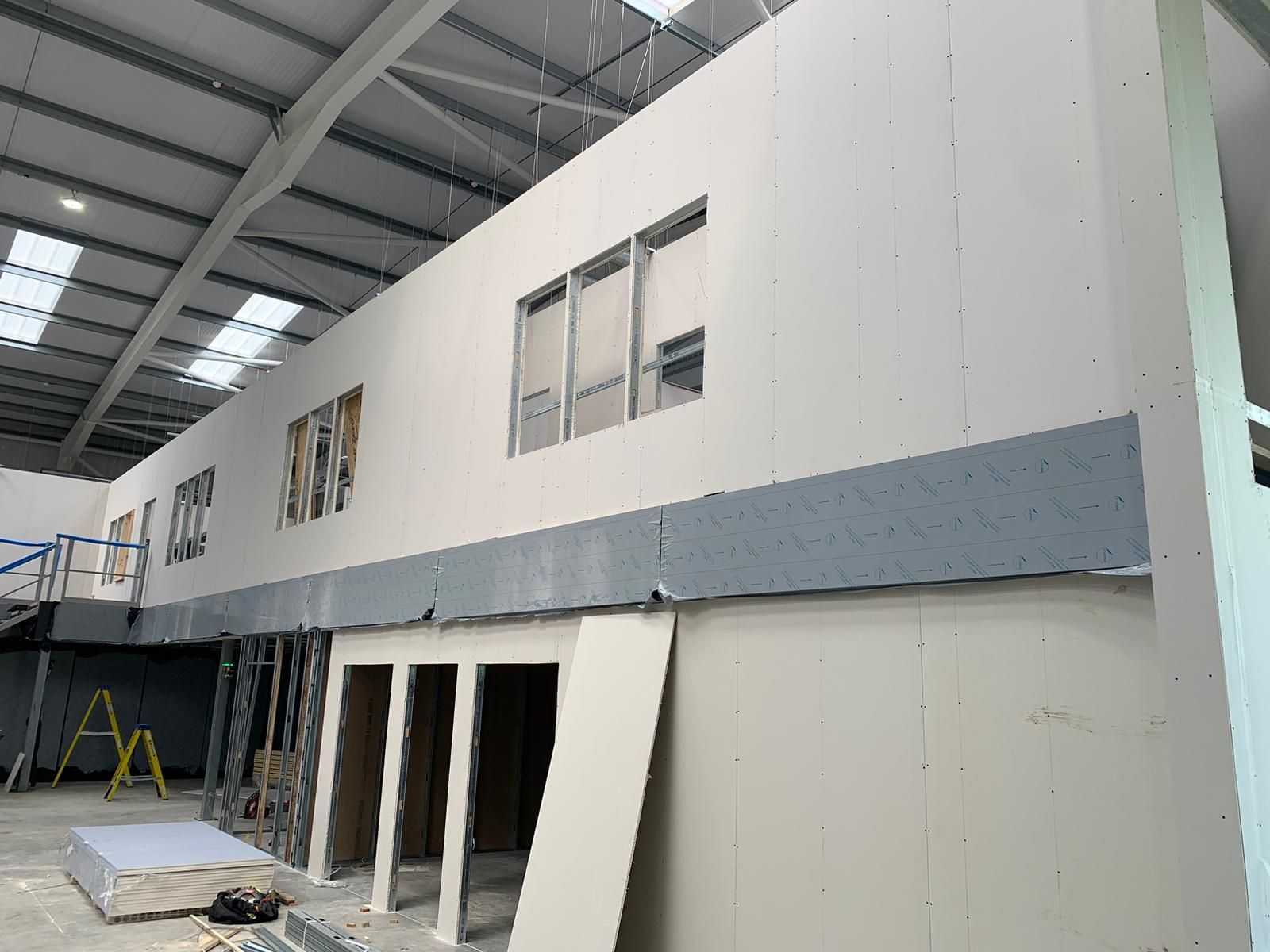 Indoor construction site with a two-story modular building, gray panels, and open doorways under a warehouse roof