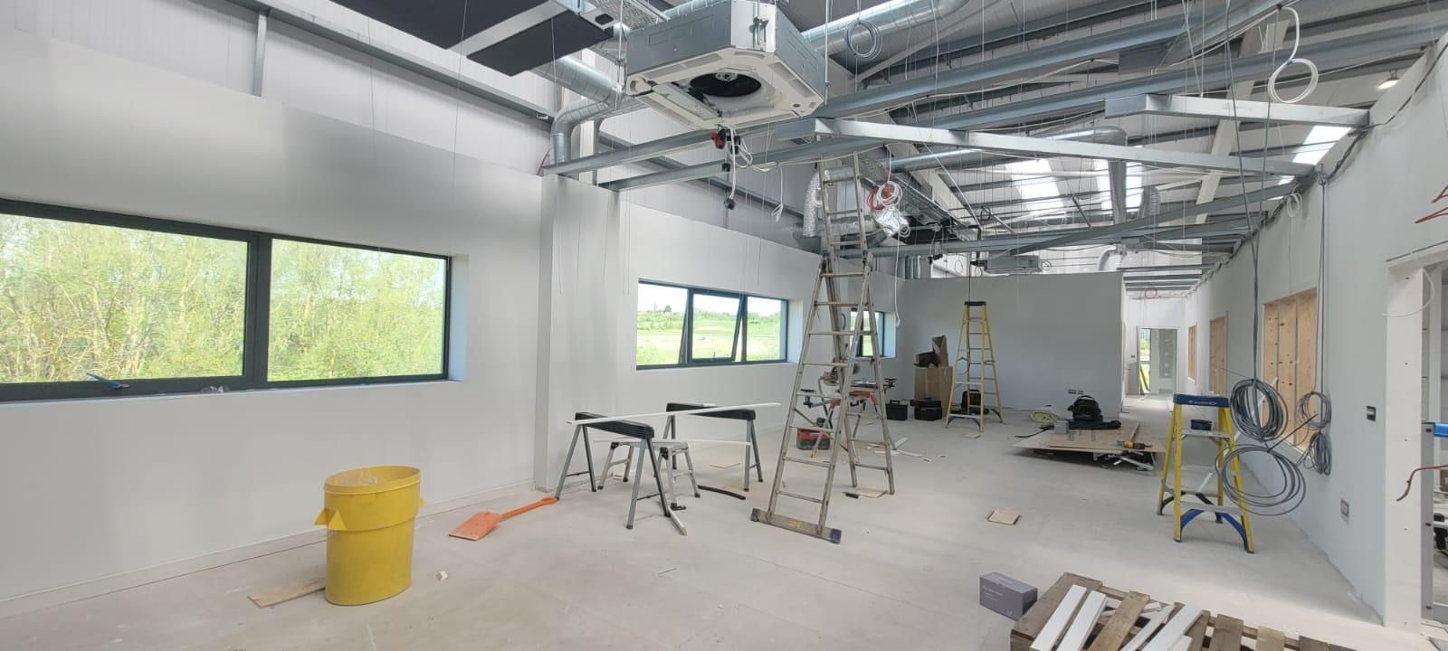 Empty office space under renovation with exposed ceiling, ladders, tools, and a yellow bin