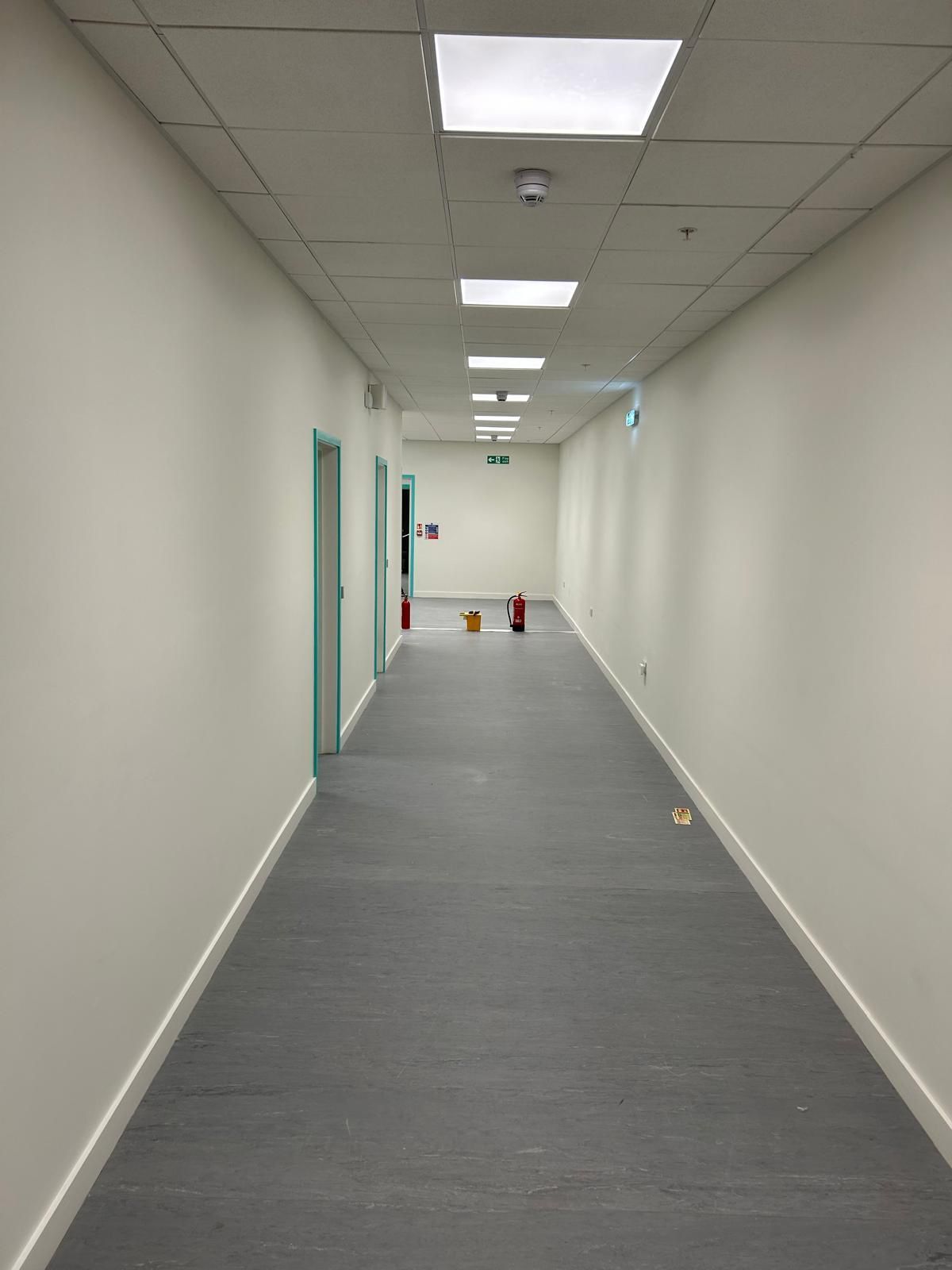Empty office hallway with white walls, gray carpet, and ceiling lights, showing a few cones in the distance